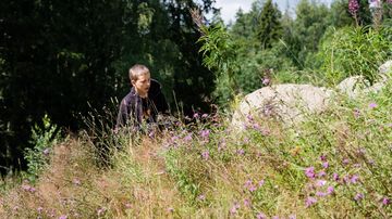 En tonårig pojke står på en äng vid en stensamling med skog i bakgrunden.