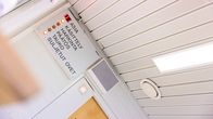 Kuvassa näkyy taulu, jossa on merkkivalot ja teksti. Teksti: ASIA, KÄSITTELY, HARKINTA, PÄÄTÖS, TAUKO, SULJETUT OVET. Näytetään, että "ASIA" on aktiivinen valolla. Taulu on kiinnitetty valkoiseen seinään katon lähellä. Vieressä on harmaa laite ja valkoinen katto, jossa on pyöreä ilmanvaihtoaukko ja suorakulmainen valo.
