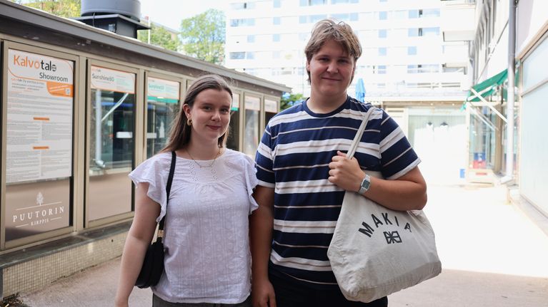Två unga personer står utomhus framför en byggnad med stora fönster och informationsskyltar.