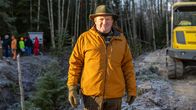 En person står i förgrunden av bilden, klädd i en gul jacka och hatt, och i bakgrunden syns en grupp människor och en grävmaskin på en skogsväg.