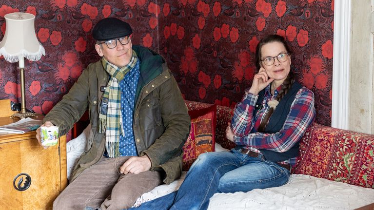 Juho och Monica sitter på en soffa i ett rum med rödblommig tapet.