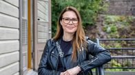 Nainen istuu puisella tuolilla ulkoterassilla vaalean rakennuksen edessä, yllään musta nahkatakki ja “Valentino”–tekstillä varustettu paita.