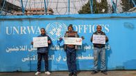 Kolme ihmistä pitelee kylttejä UNRWA:n toimiston portin edessä.