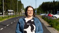 Nainen seisoo autotien vieressä pukeutuneena denim-takkiin ja kukkakuosiseen mekkoon. Taustalla näkyy autojen parkkipaikka ja sähkölinja, sekä kesäinen metsänreuna.