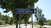 Tienviitta, jossa teksti "Variskan koulu kirjasto bibliotek", vihreitä puita ja tyhjä katu taustalla.
