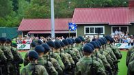 Militärparad med rekryter ute på en gräsplan. I bakgrunden pubilk.