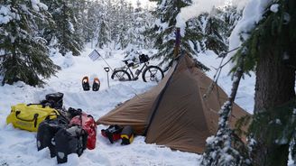 Talvipyöräretkeilyä Lapissa: Polkupyörä nojaa puuhun lumen peittämässä metsässä, jossa on pystytetty teltta. Teltan edustalla on retkeilyvarusteita ja lumikenkäpareja, ja kamiinapiipusta nousee savua.
