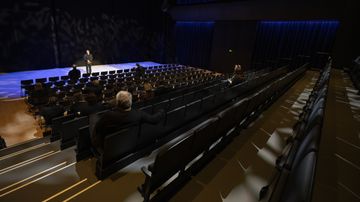 En person står på en scen inför gles publik i en mörk sal med många tomma stolsrader.