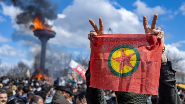 En person i en stor folksamling håller Kurdiska arbetarpartiets flagga och visar segertecken. I bakgrunden brinner eldar.