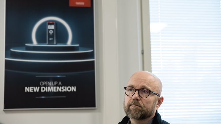 Porträttfoto på en person i bilden som bär mörka glasögon. I bakgrunden syns ett fönster med persienner och en affisch med texten "Open up a new dimension".