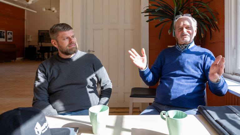 Mathias Blomqvist och Christer Westerholm sitter vid ett bord i ett solbelyst rum med orange väggar.