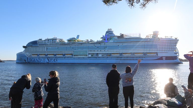 A large white cruise ship sails past a group of around ten people on the shoreline who are waving on a sunny day.