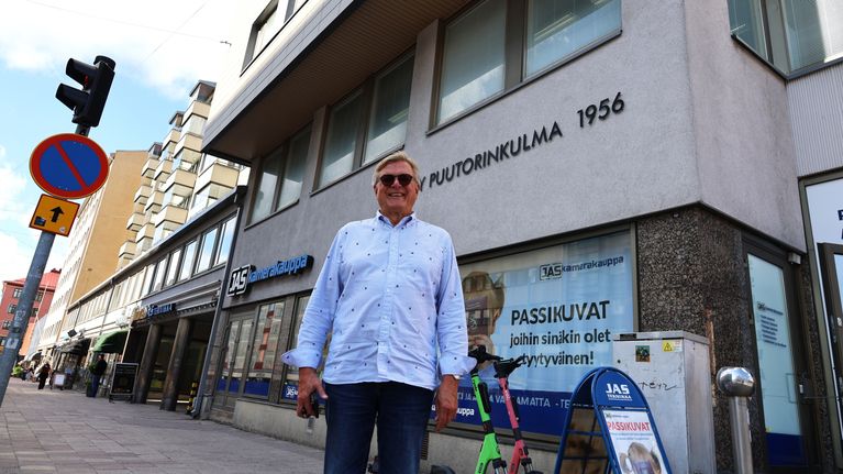 En man står på trottoaren framför en byggnad med skylten "PUUTORINKULMA 1956" och flera butiksskyltar.