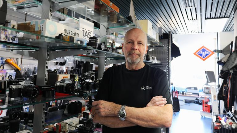 En man med skägg står i en butik framför hyllor med kamerautrustning och fototillbehör.