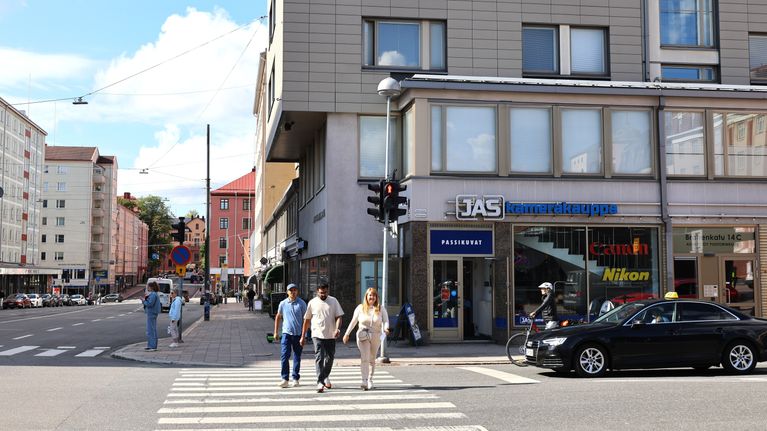 Tre personer går över ett övergångsställe framför en byggnad med skylten "JAS fotohandelgrupp" och en parkerad bil.