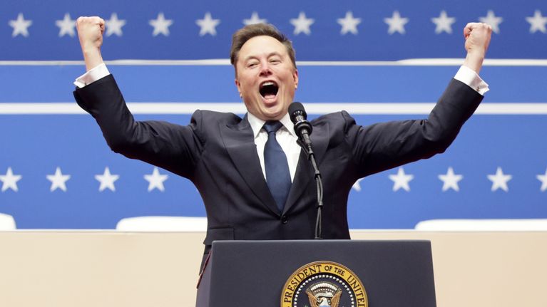 Elon Musk seisoo puhujankorokkeella ja elehtii kädet ylös nostettuina ja huutaa yleisölle.