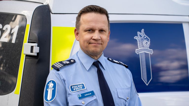 En polis i ljusblå uniform står framför ett polismärke och en bil.