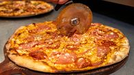 En hand håller en pizzaskärare och skär upp en gräddad pizza med ost, skinka och pepperoniskivor på en träbricka.