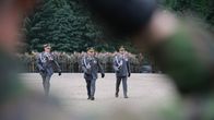 Kolme juhlapukuista upseeria kävelee aukiolla, taustalla seisoo rivissä sotilaita.