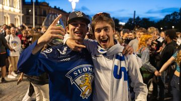 Kaksi nuorta miestä Suomen faniasusteissa tuulettaa iloisena kameralle. Taustalla täysi tori.