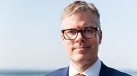 Silmälasipäinen mies katsoo kameraan ja hänen takanaan aukeaa sumea merimaisema.