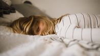 Woman lying on a bed with hair covering most of her face, but one of her eyes is looking into the camera.