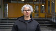 En person med lockigt hår står utomhus framför en byggnad med gula dörrar och fönster i lätt regn.