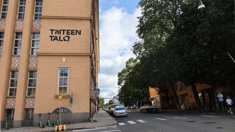 Fasad av en beige byggnad med texten "Taiteen Talo" i svart, blå himmel syns i bakgrunden.