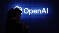 OpenAI:n logo tummalla monitoriseinämällä. Kaksi hahmoa sen edessä näprää kännyköitä.