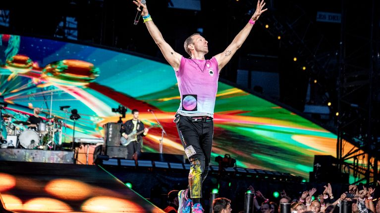 Chris Martin laulaa mikkiin lavalla. Taustalla näkyy Parken stadionin rakenteita ja yleisön muodostama valomeri.