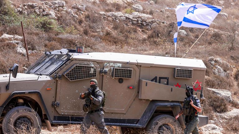 Kaksi aseistettua sotilasta kävelee panssaroidun ajoneuvon edessä, jonka päällä on Israelin lippu.