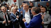 Useita ihmisiä seisoo sisätiloissa lähekkäin, osa hymyilee ja keskustelee, taustalla näkyy televisiokameroita ja mikrofoneja.