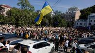Suuri joukko ihmisiä seisoo kadulla autojen ympärillä, yksi henkilö pitelee Ukrainan lippua.