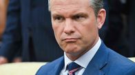 Pete Hegseth istuu vaalealla sohvalla tummansinisessä puvussa ja raidallisessa solmiossa, taustalla näkyy ihmisiä.