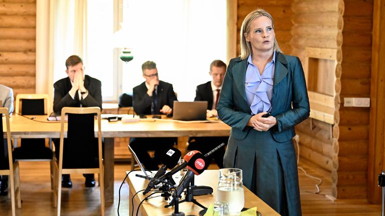 Riikka Purra seisoo yleisön edessä ja katsoo seinällä olevaa esitystä. Taustalla istuu miehiä.