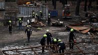 Palomiehiä ja työntekijöitä raivaustöissä palaneen rakennuksen jäännöksissä, taustalla kaivinkone ja roskalavoja.