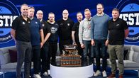 Kahdeksan miestä seisoo studiossa pöydän äärellä, jonka päällä on suuri jääkiekkopokaali. Taustalla näkyy teksti “MESTIS”.