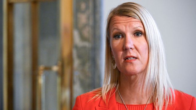 Vaaleahiuksinen nainen punaisessa jakussa seisoo sisätilassa.