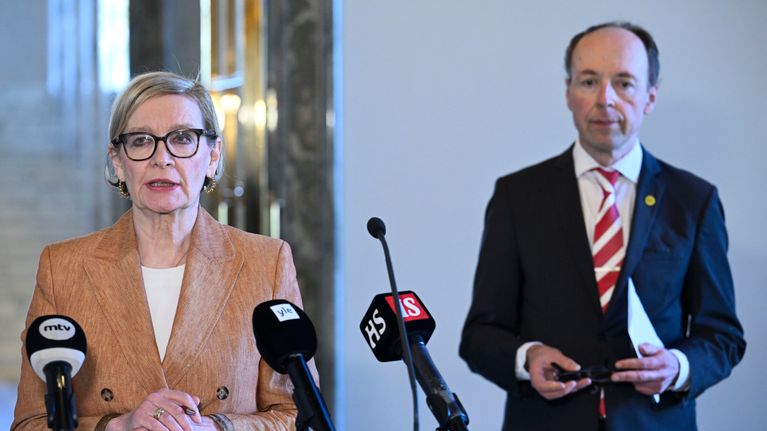 Kaksi ihmistä seisoo puhujakorokkeen äärellä mikrofonien edessä, taustalla portaikkoa ja heijastavia pintoja.