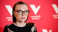  Minja Koskela seisoo punaisen taustan edessä, jossa valkoinen V-kirjain ja teksti "vasemmistoliitto".