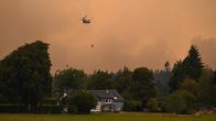 Helikopteri lentää savuisella taivaalla vesivaijerin kanssa omakotitalon ja metsän yläpuolella.