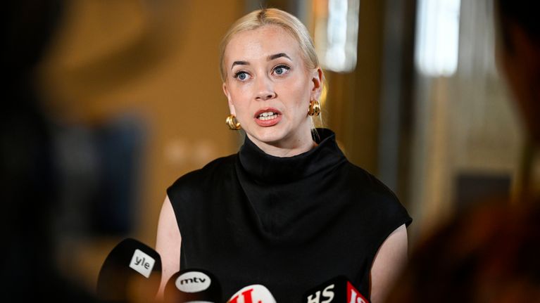 A blonde woman stands at a podium, speaking into microphones. 