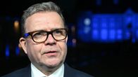 Närbild av Petteri Orpo i en mörkblå kostym. Han har glasögon på sig och tittar framåt.
