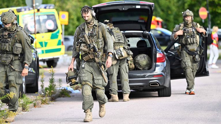 Tungt beväpnade personer i kamouflagekläder röd sig nära sin bil, som har bagageluckan öppen. En bär sin hjälm i handen.