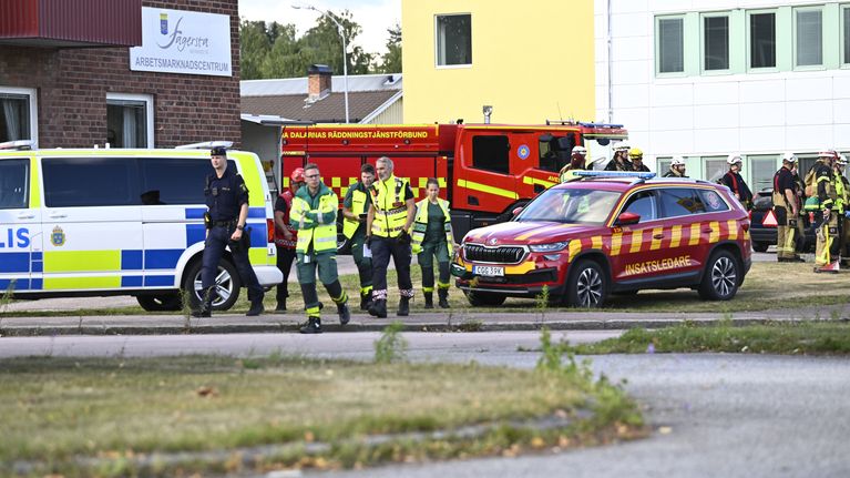 Useita poliiseja ja pelastushenkilöitä seisoo ajoneuvojen edessä rakennusten läheisyydessä, taustalla paloauto.