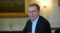 Petteri Orpo istuu sisätiloissa mustassa puvussa vaalean paidan kanssa, taustalla suuri maisemamaalaus.