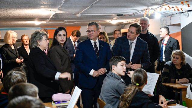 Useita aikuisia seisoo luokkahuoneessa, jossa istuu lapsia pulpeteissa; taustalla syksyisiä koristeita katossa.