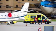 En ambulans står parkerad vid ett mindre flygplan på en flygplats, omgiven av flera personer i arbetskläder.