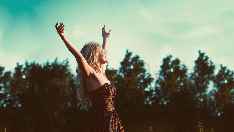 Woman on a sunny day wearing a long dress walking through a field and stretching her arms up in the air.
