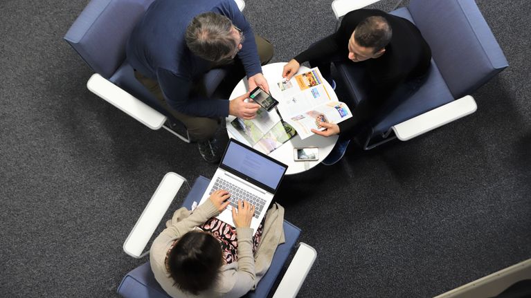 Tre personer sitter runt ett litet bord och arbetar med dokument och en bärbar dator.